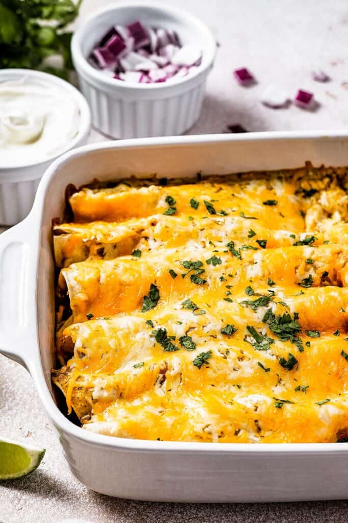 Baked chicken enchiladas with a dish of sour cream and a dish of chopped red onion.