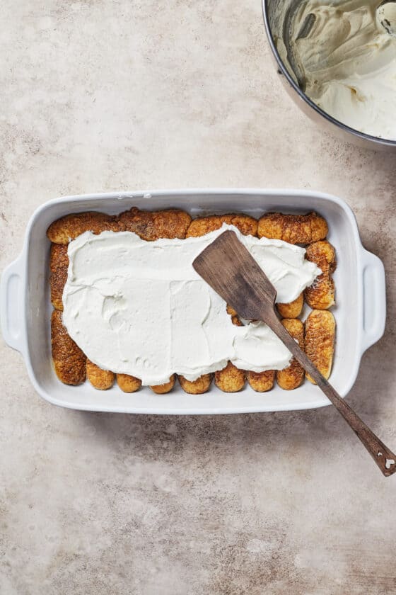 Creamy topping partially spread across a layer of savoiardi in a baking dish. A metal spatula is in the dish as well.