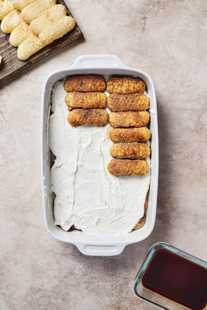 A baking dish with a layer of cookies being added to layers of cookies and mascarpone cream.
