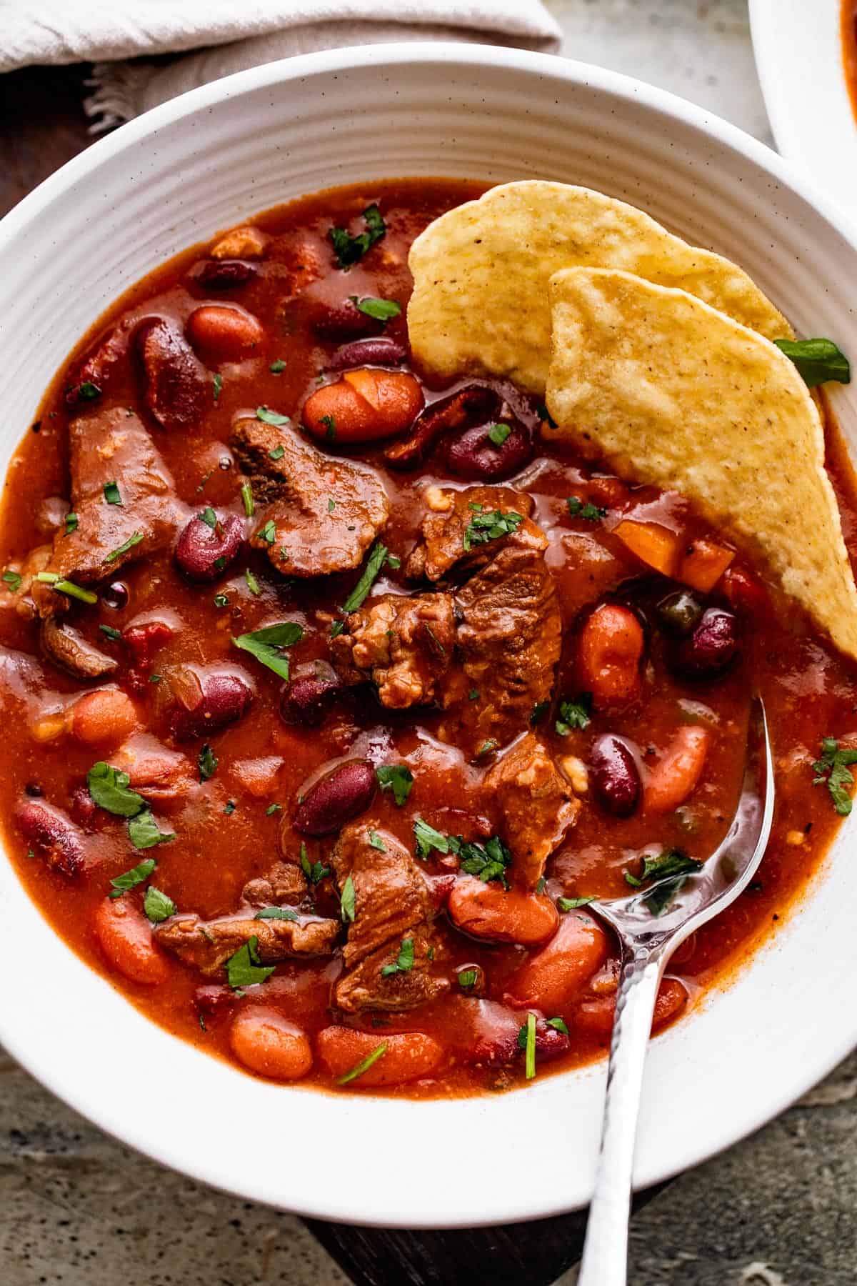 overhead shot of chili con carne in a white bowl, with a spoon and tortilla chips set inside the chili