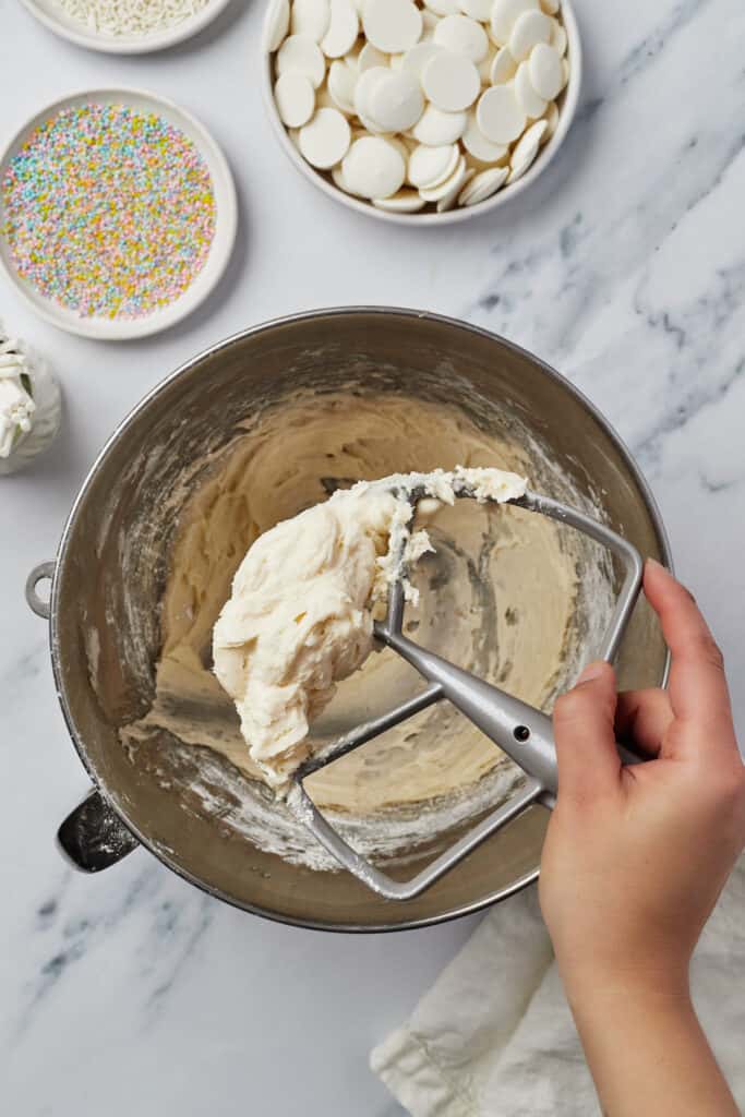 Frosting in a mixing bowl. A woman's hand is lifting a stand mixer attachment covered with frosting out of the bowl.