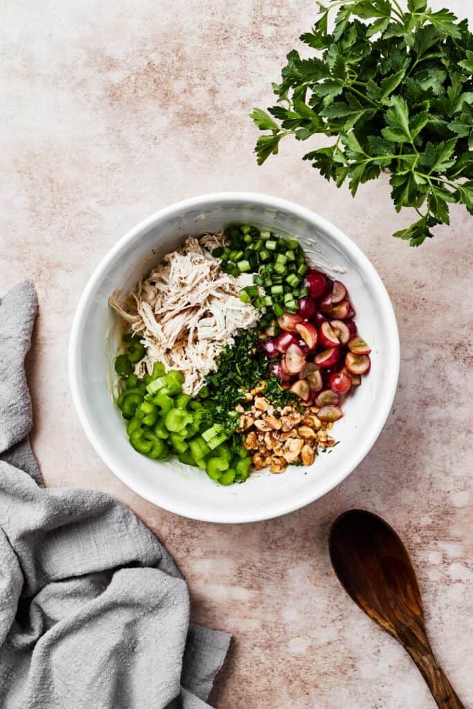A white mixing bowl with shredded chicken, vegetables, grapes, and other ingredients.