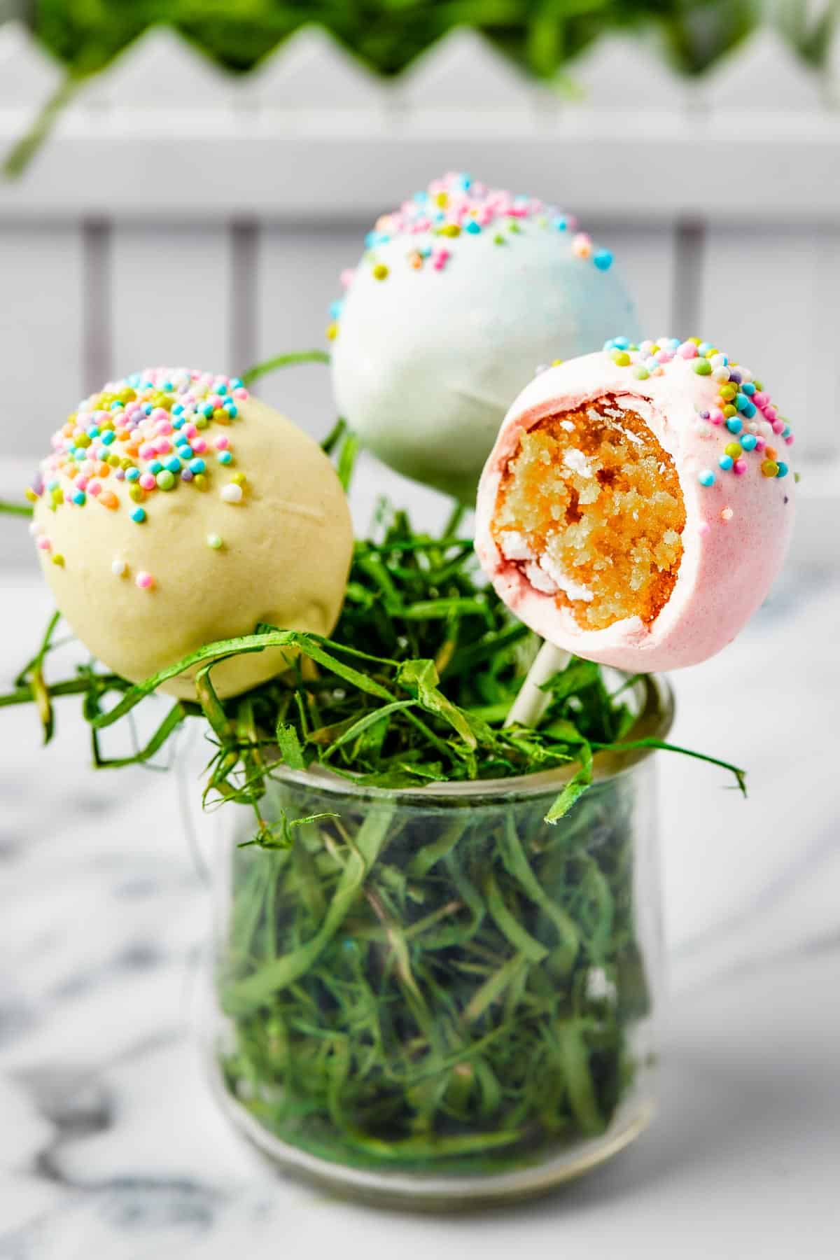 Three cake lollipops in a small glass filled with Easter grass. One of the lollipops has had a bite taken out of it.