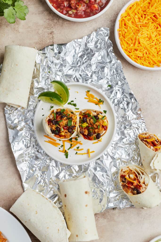 A plate with two burrito halves on it, garnished with cheese and lime wedges. Aluminum foil, more burritos, and a dish of shredded cheese are placed around the plate.