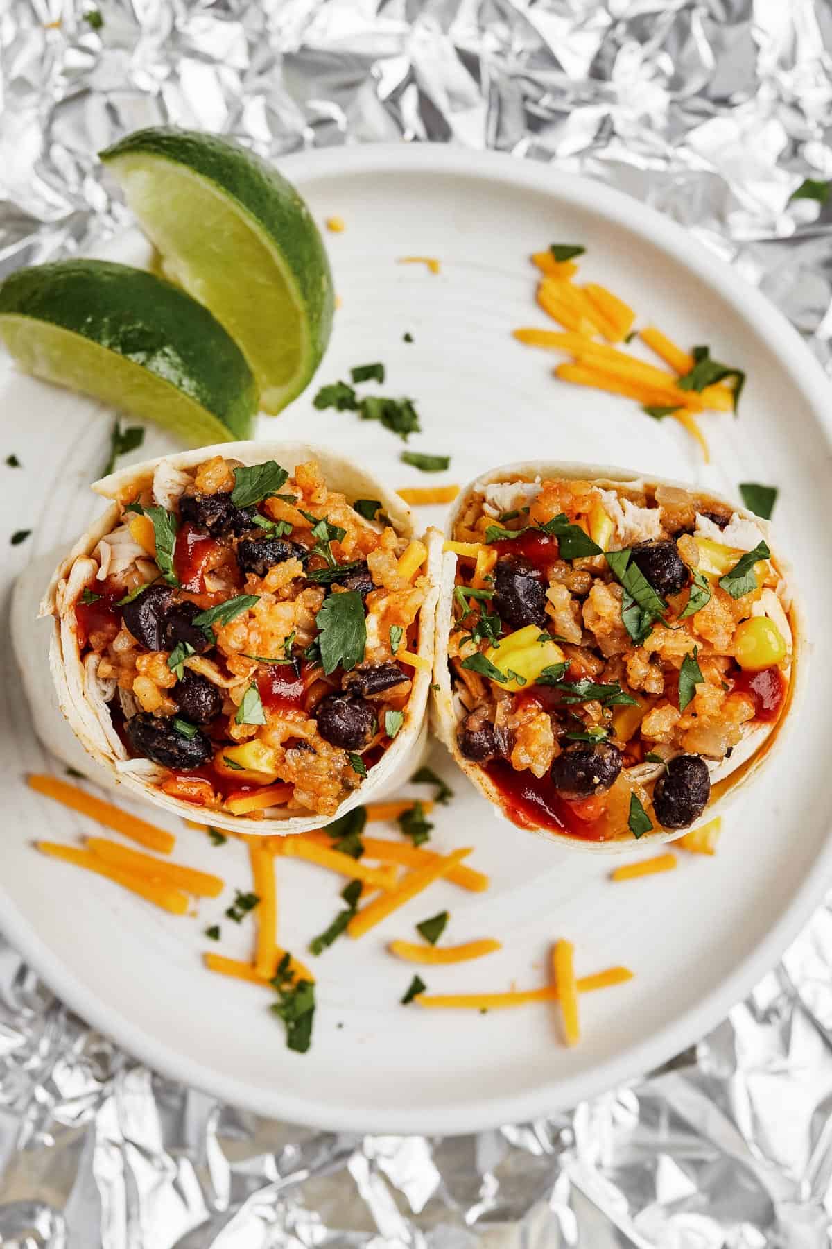 A plate with two burrito halves on it, garnished with cheese and lime wedges. Aluminum foil, more burritos, and a dish of shredded cheese are placed around the plate.