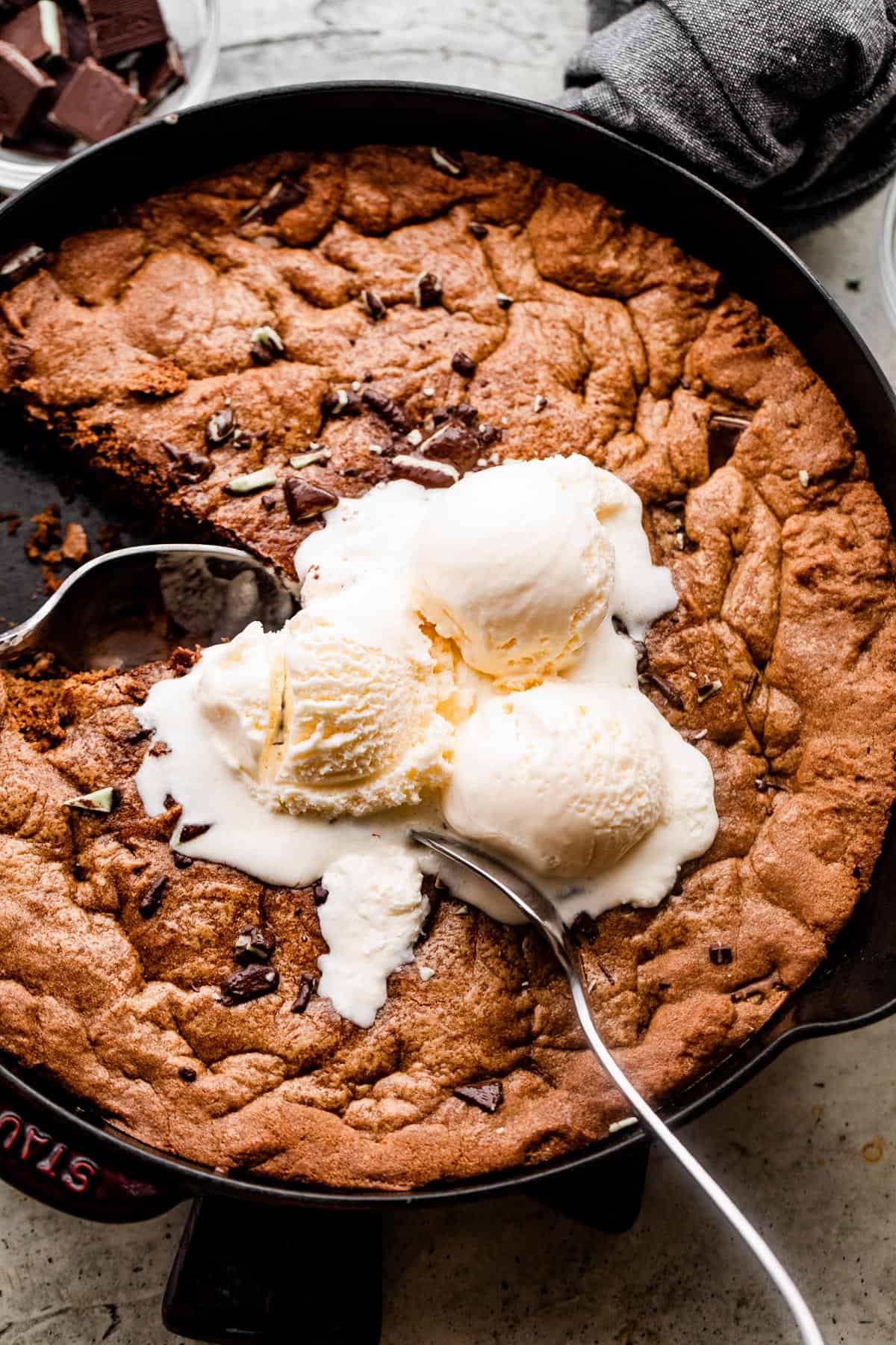 overhead shot of a skillet cookie inside the skillet, with three vanilla ice cream scoops on top and two large spoons digging into the cookie.