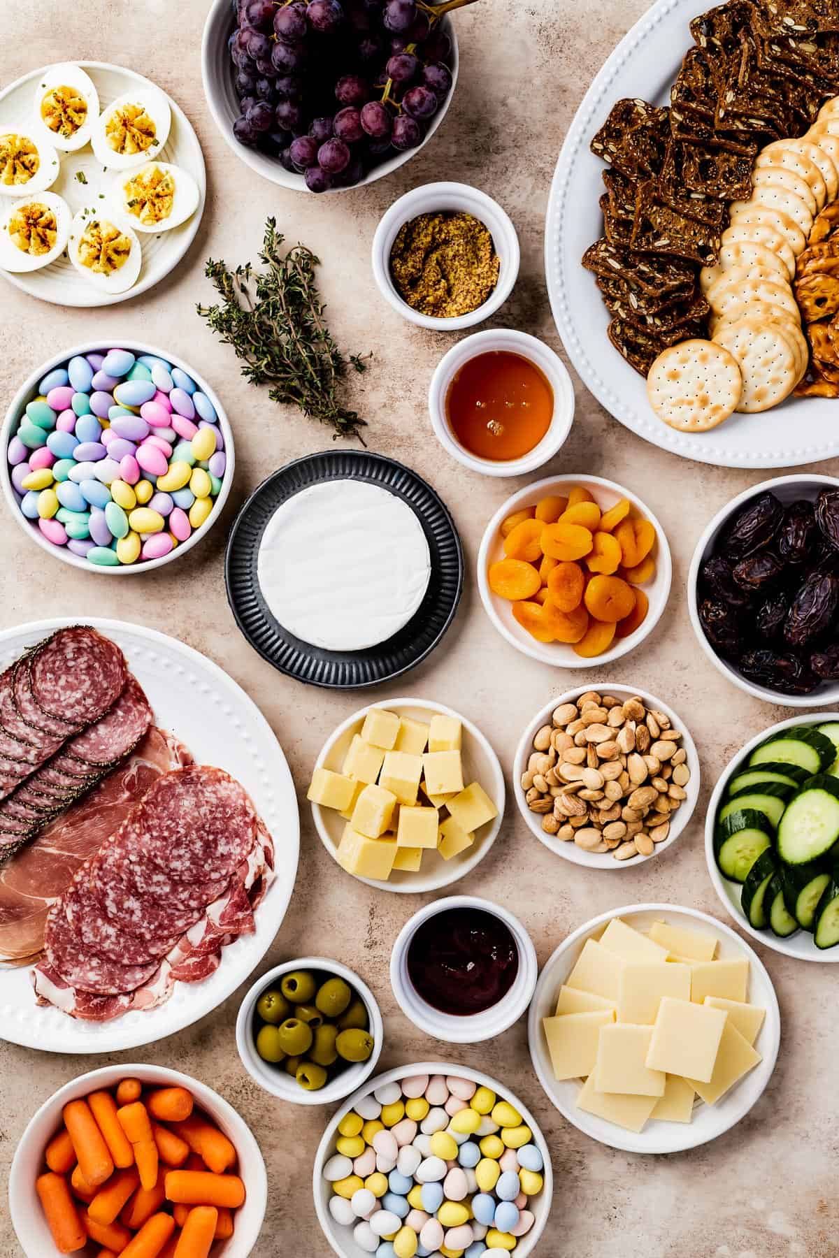 From top left: Deviled eggs, grapes, a platter of crackers, jordan almonds, fresh thyme, grainy mustard, strawberry preserves, a wheel of brie, dried apricots, medjool dates, a plate of sliced meats, cubed white cheddar, marcona almonds, sliced cucumber, castelvetrano olives, honey, gouda.