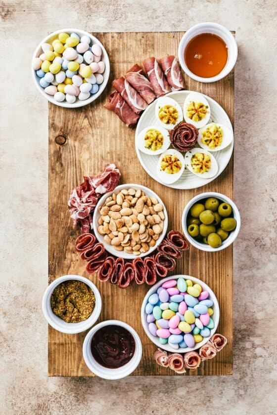 A rectangular cutting board with bowls of olives, condiments, jordan almonds, marcona almonds, and a plate of deviled eggs placed at random.