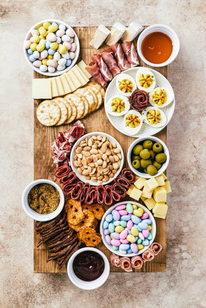 A wooden cutting board with dishes of ingredients, folded and rolled sliced meats, sliced and cued cheeses, and an assortment of crackers arranged on top.
