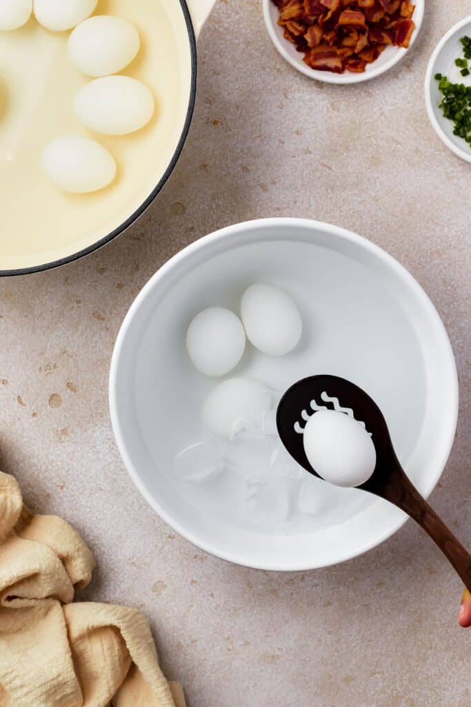 A bowl of peeled hard cooked eggs in water. Nearby are a plate of eggs in the shell, and a small dish of discarded egg shells, along with a few small dishes of ingredients and a cloth napkin.