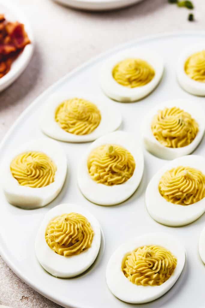 A shallow white platter, shaped like an egg, with deviled eggs arranged on it. Bowls of garnishes are arranged around the platter.