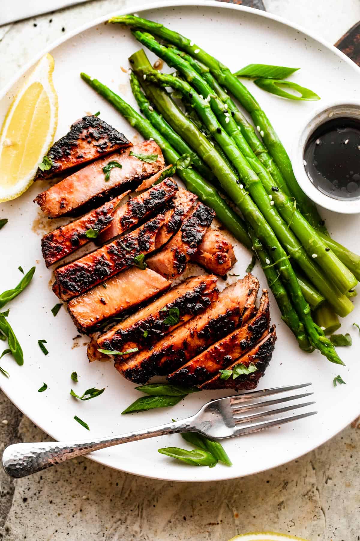 Overhead shot of a sliced tuna steak served on a white dinner plate with asparagus stalks, dipping sauce, and lemons.