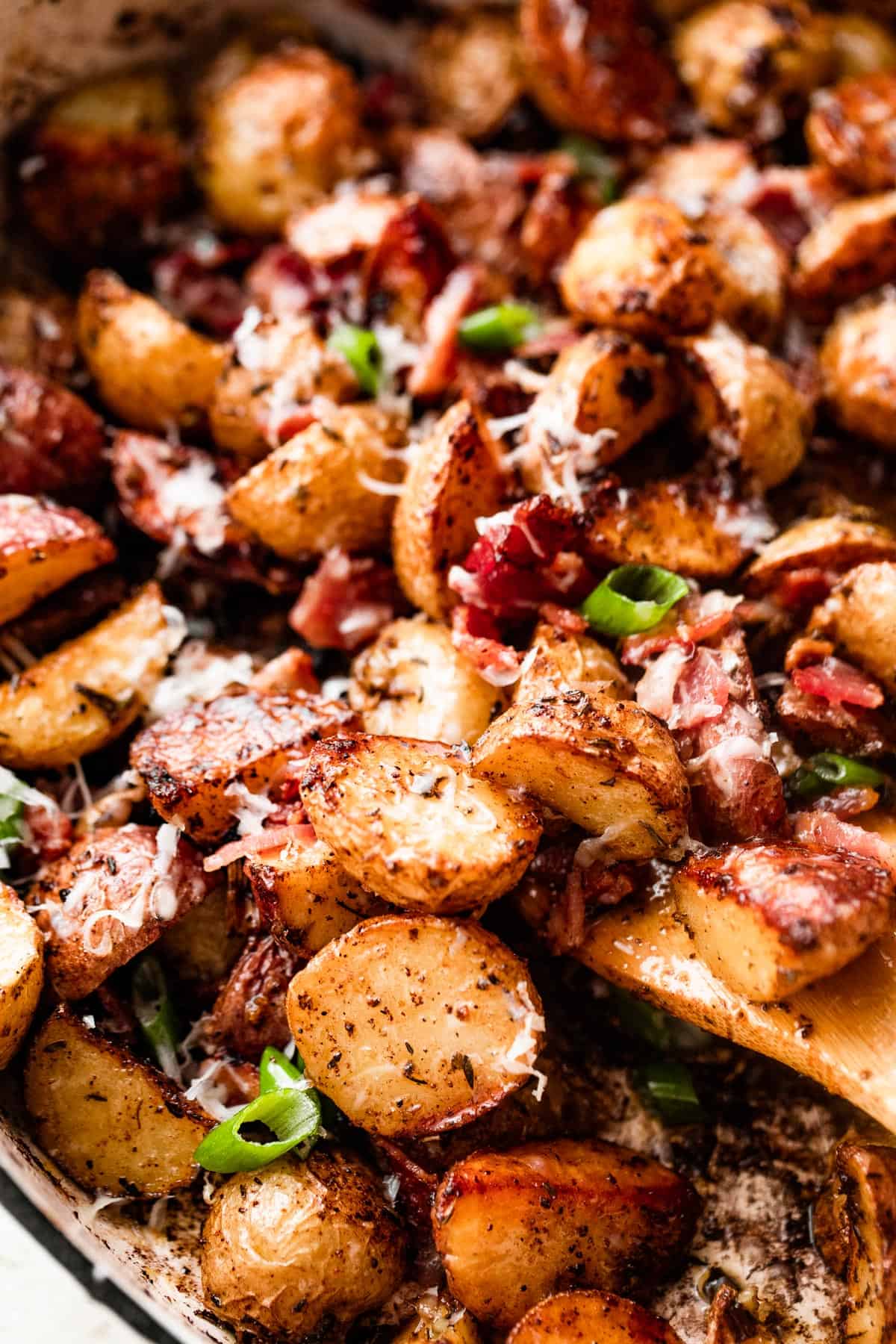 close up shot of a wooden spoon stirring through roasted potatoes with bacon and cheese.