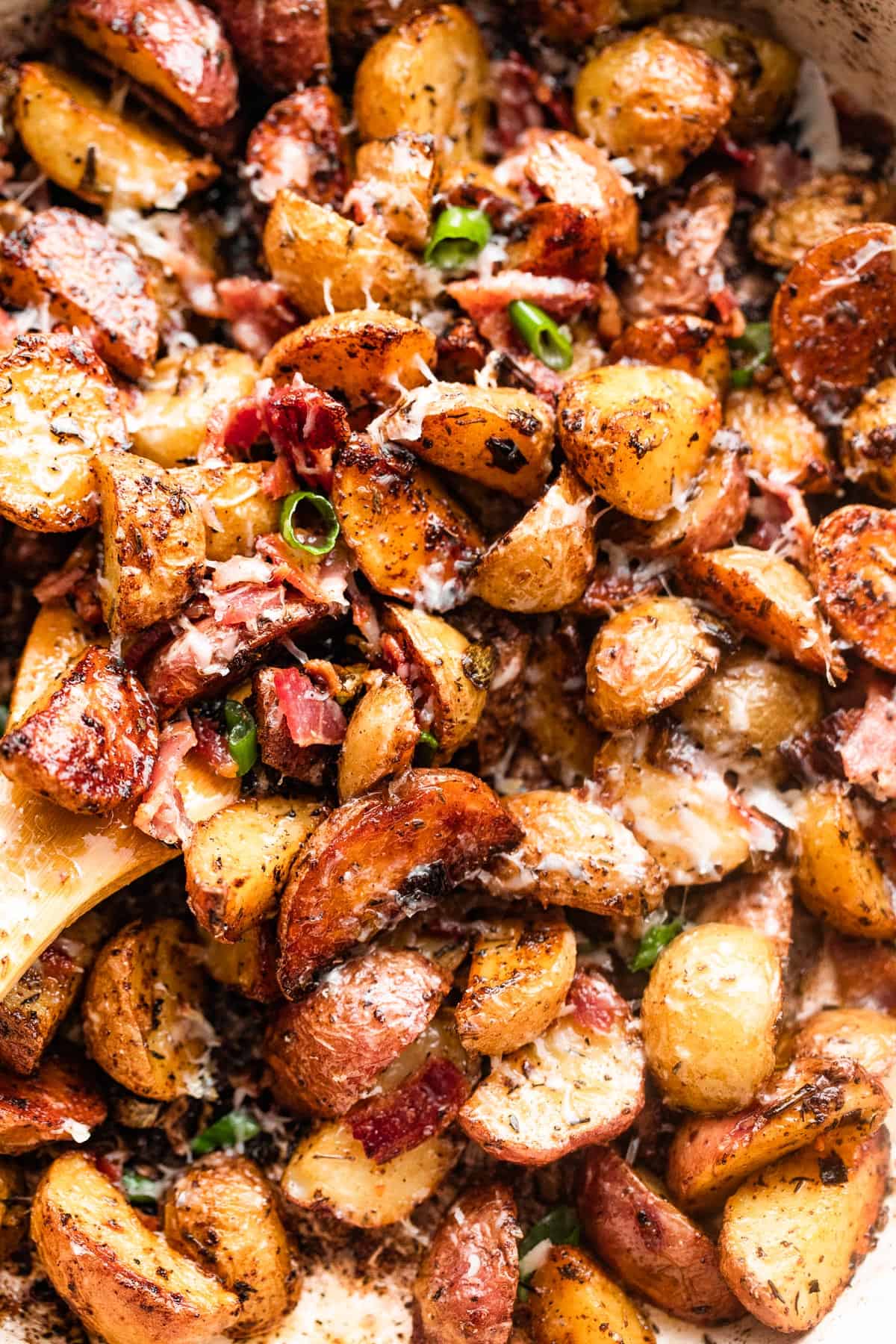 close up shot of a wooden spoon stirring through roasted potatoes with bacon and cheese.