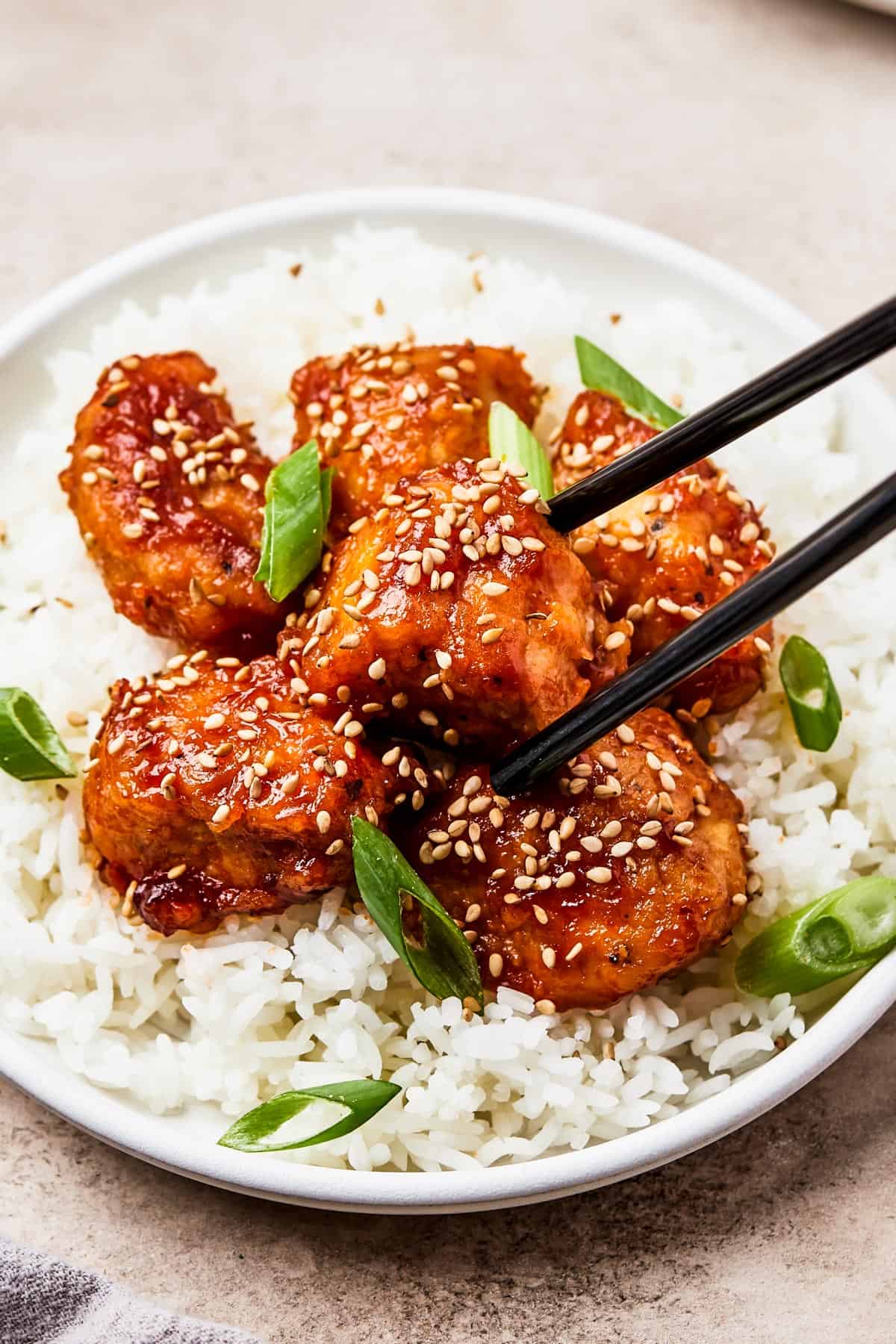 A small plate of takeout-style chicken over rice, with chopsticks holding one piece of chicken.