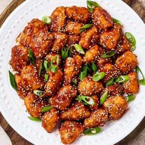 overhead shot of sweet and sour chicken served on a white plate with rice, green onions, and toasted sesame seeds.
