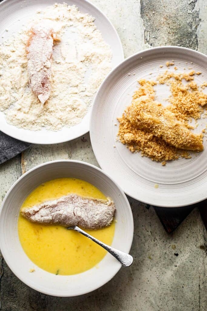 bottom left: raw chicken tender soaking in a bowl with whisked eggs. upper left: raw chicken tender in a bowl with all purpose flour. right side: raw chicken tender in pork rind breading.