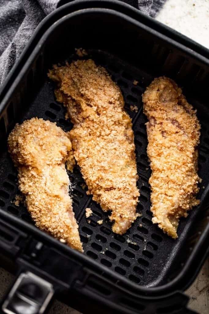 three raw breaded chicken tenders arranged inside a black air fryer basket.