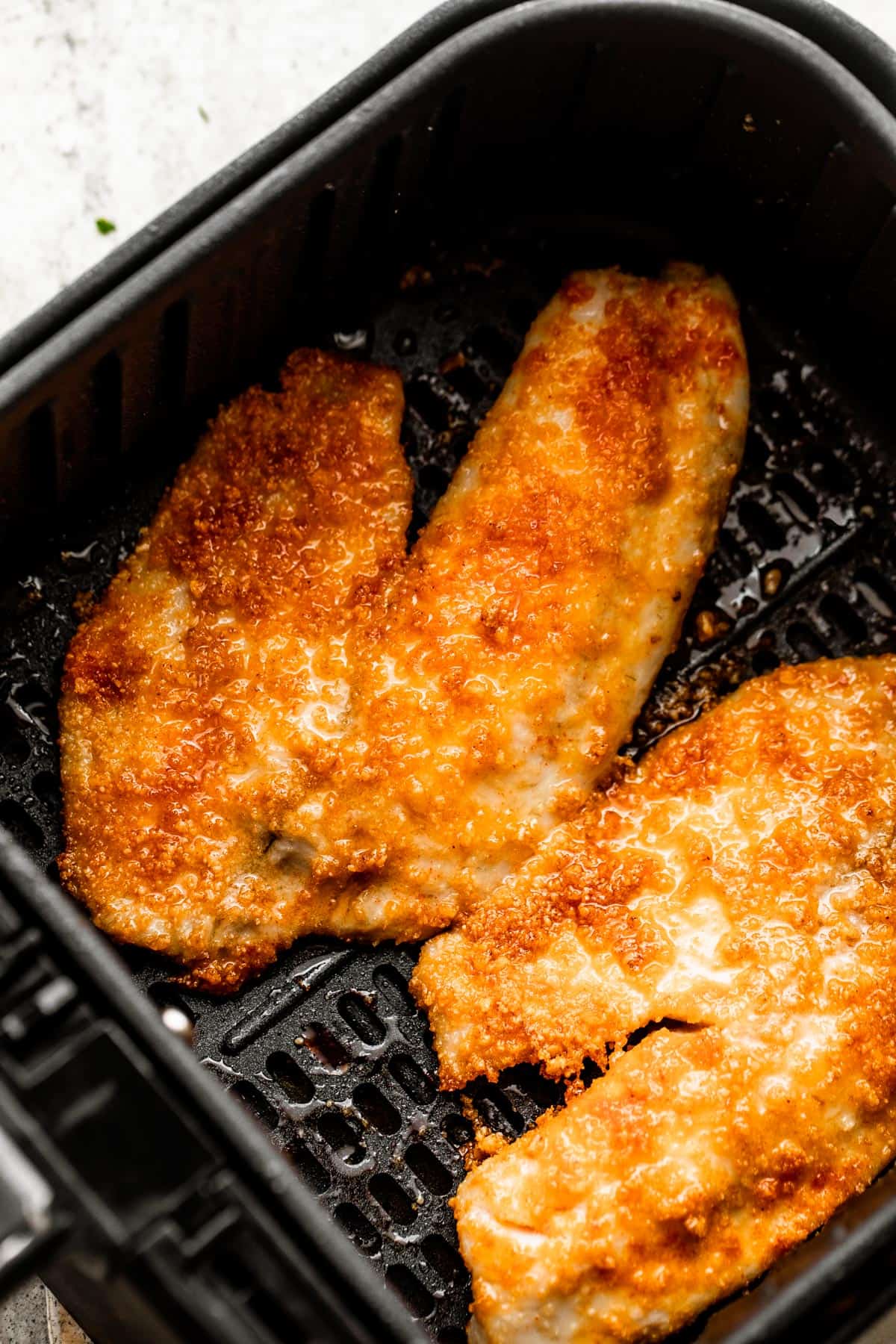 two cooked tilapia fillets arranged inside of a black air fryer basket.