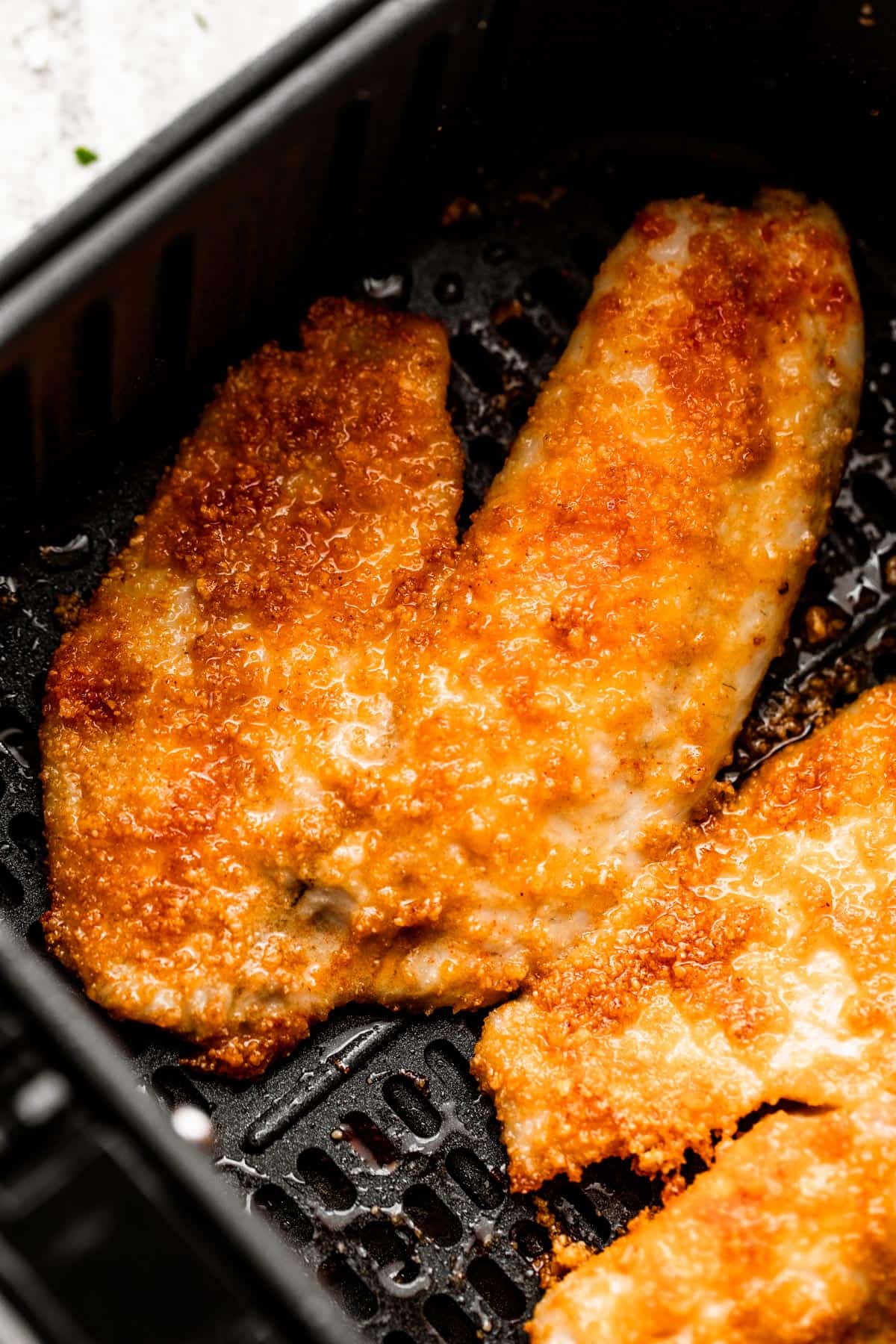 two cooked tilapia fillets arranged inside of a black air fryer basket.