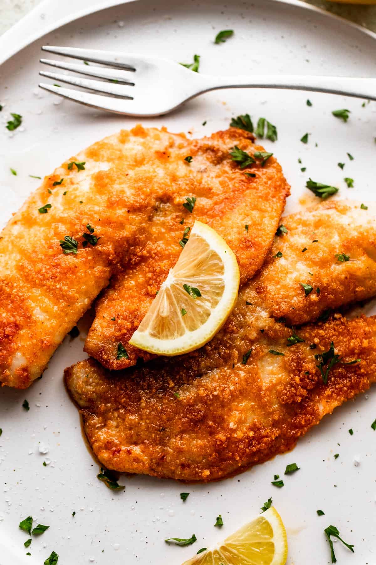 two cooked tilapia fillets placed on a white plate and topped with a slice of lemon.