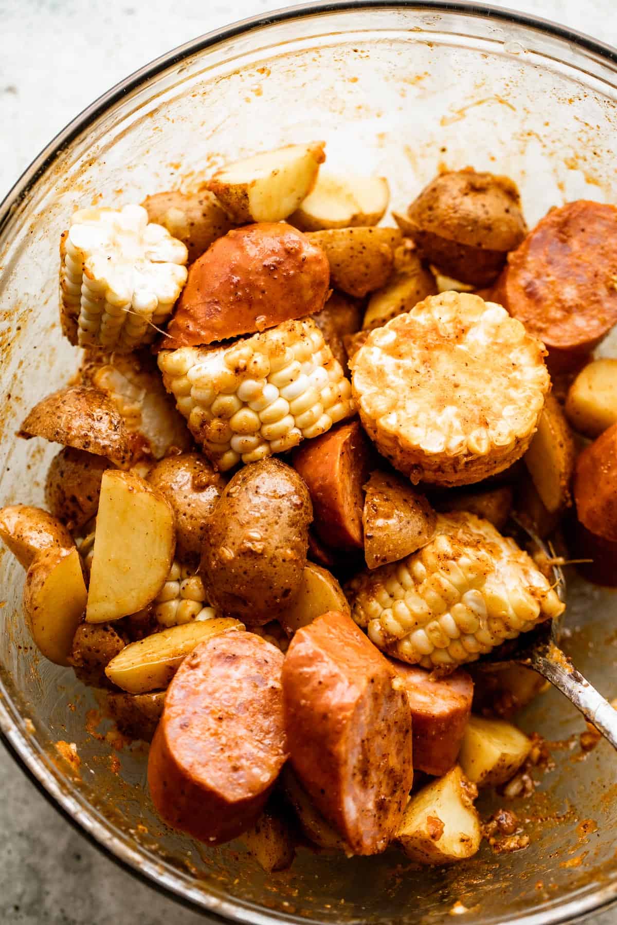 overhead shot of sliced potatoes, corn on the cob rounds, andouille sausage rounds, and shrimp in a glass bowl.