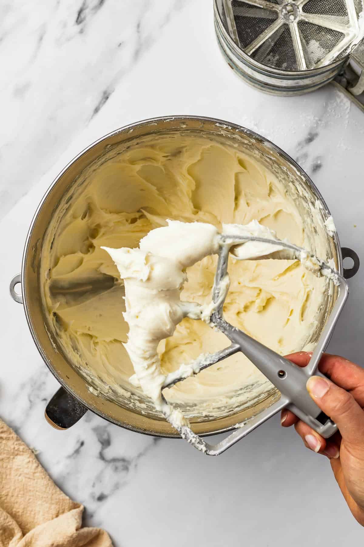 A metal mixing bowl with whipped frosting inside. A woman's hand is lifting a beater attachment out of the frosting.