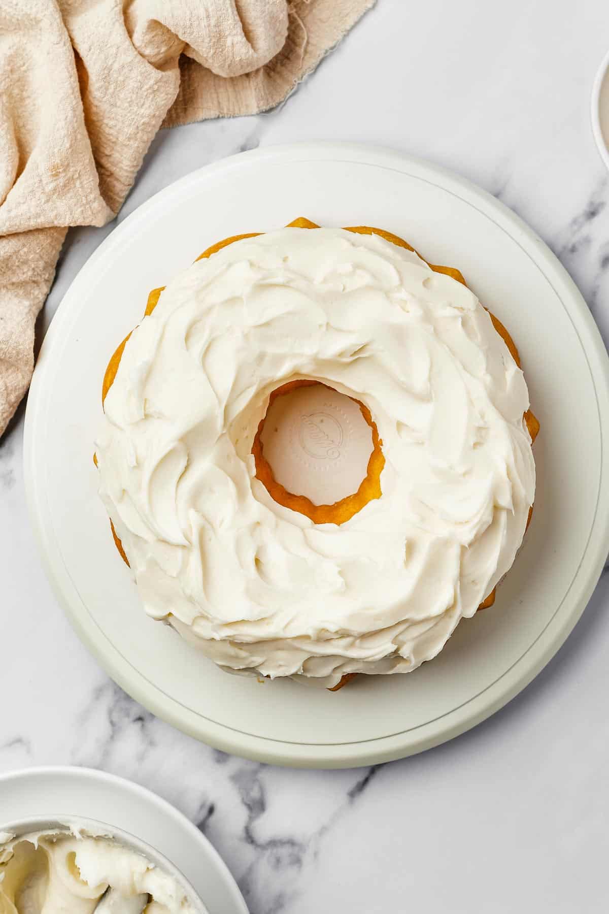 Overhead shot of a bundt cake iced with thick, creamy frosting. A bowl of frosting and dish of lemon zest are nearby on the table.