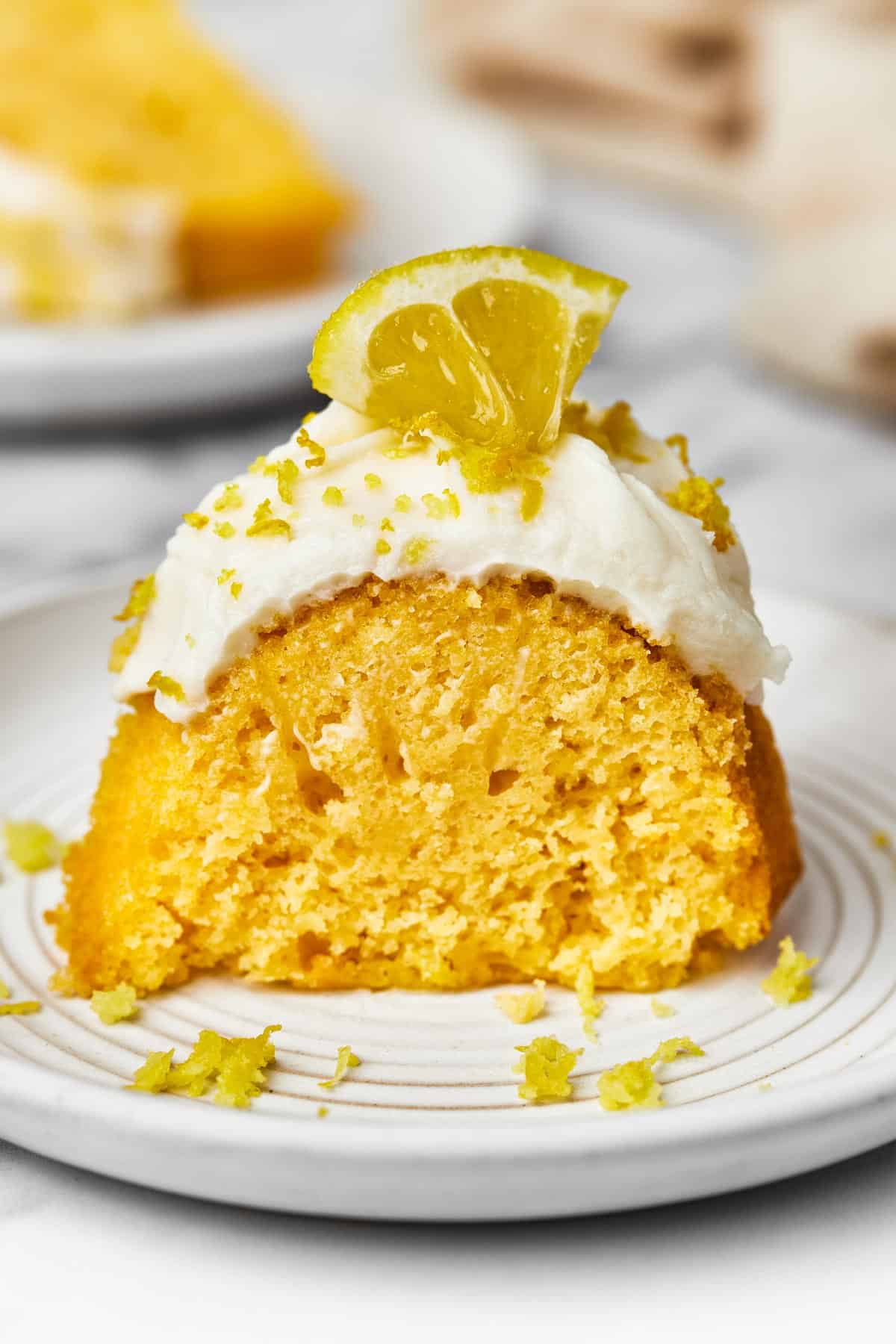 A slice of frosted cake, garnished with lemon zest and a small lemon wedge.