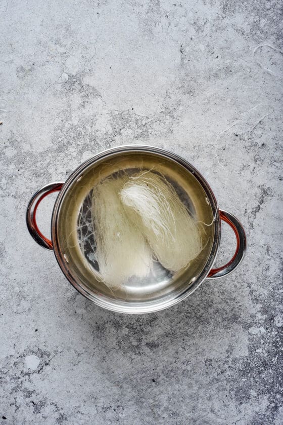 Glass noodles rehydrating in a stainless steel pot of hot water.