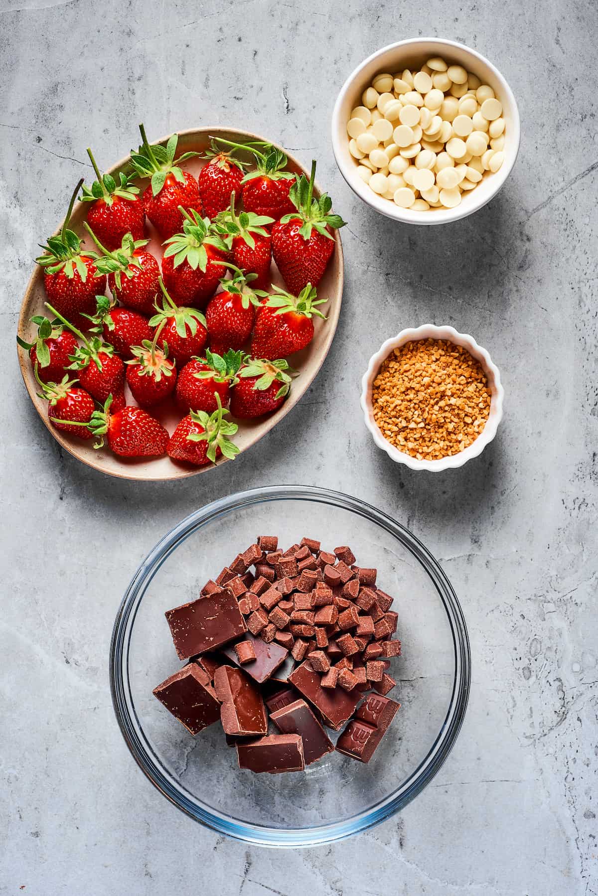 From top left: Fresh strawberries with stems, white chocolate chips, toasted chopped hazelnuts, and bowl with chopped semisweet chocolate and milk chocolate chips.