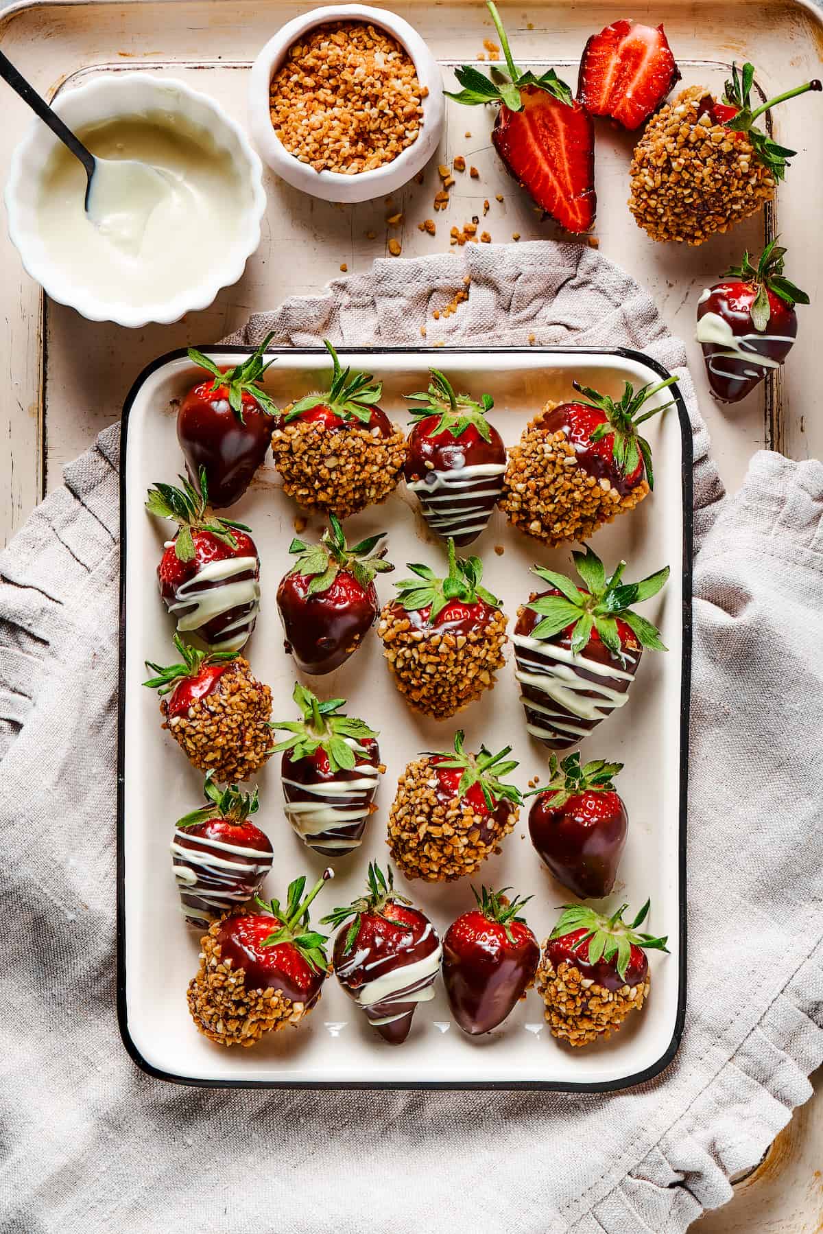 Chocolate covered strawberries on a tray, set on a decorative cloth dishtowel next to a bowl of white chocolate.