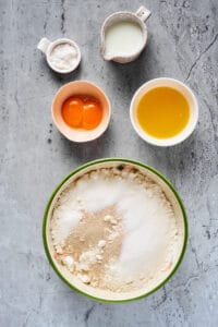 Dry baking ingredients mixed in a large mixing bowl. Sugar, milk, egg yolks, and melted butter are near the bowl in separate dishes.