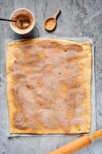 A work surface with a large rectangle of dough, a rolling pin, and a small bowl of cinnamon sugar nearby. Cinnamon sugar has been sprinkled generously all over the dough.