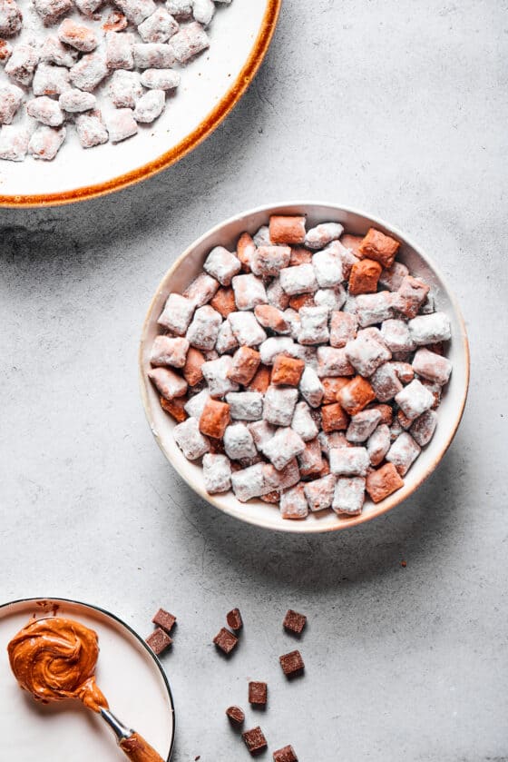 Muddy buddies in a white bowl, with a spoonful of peanut butter and a larger serving dish of the snack nearby.