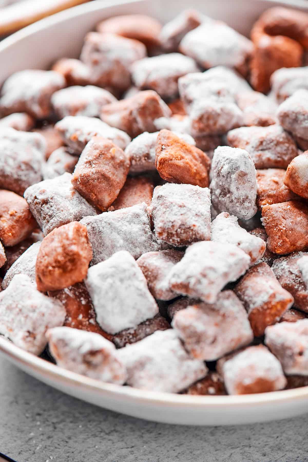 Close-up shot of puppy chow in a white bowl, to show the texture. Some pieces are fully coated in powdered sugar, while others are not.