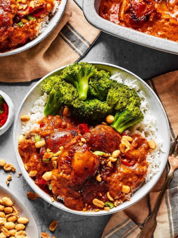 Peanut chicken served with broccoli over rice. A baking dish of the chicken is nearby on the table, along with garnishes and more plates.