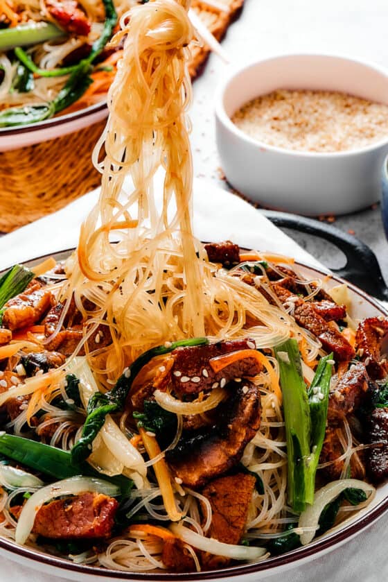 A bowl of beef stir fry with glass noodles. Some of the noodles are being lifted from the bowl with chopsticks.
