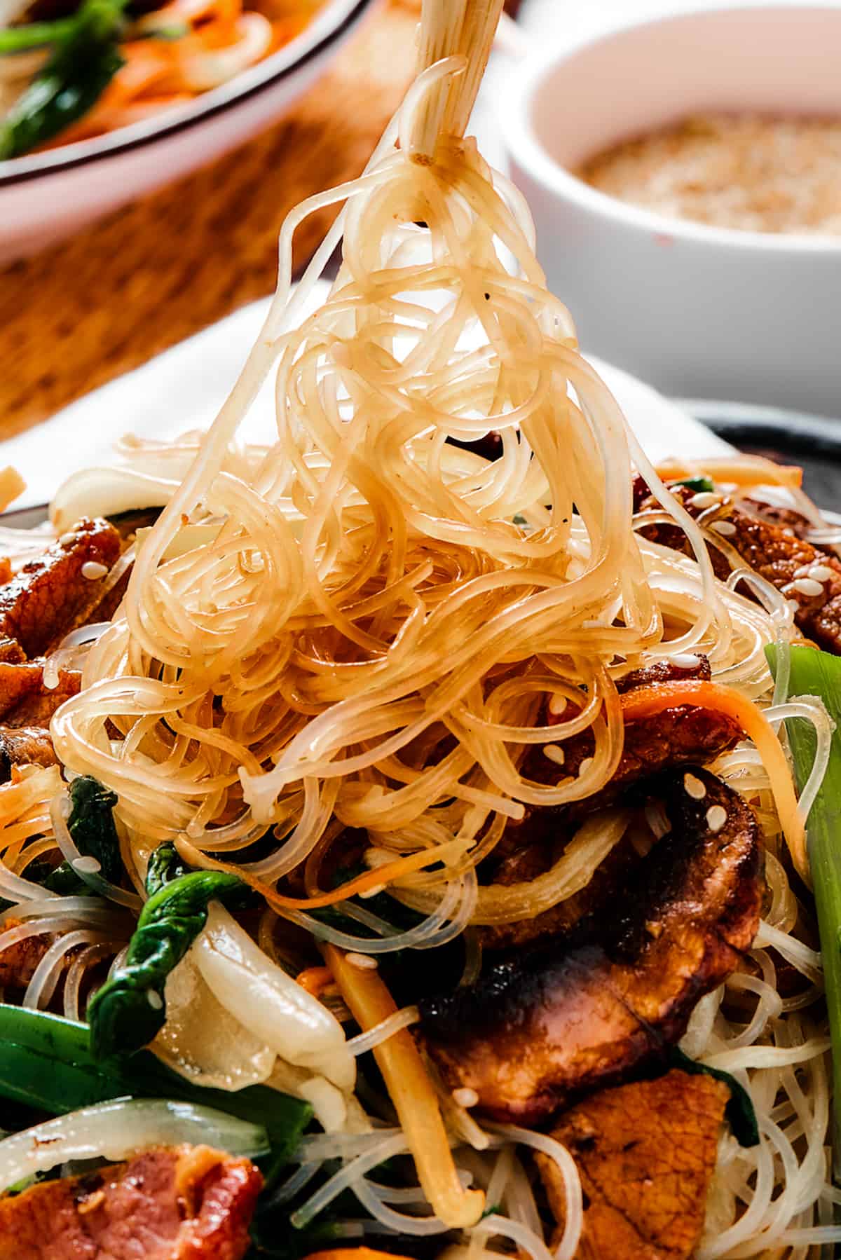 A bowl of beef stir fry with glass noodles. Some of the noodles are being lifted from the bowl with chopsticks.