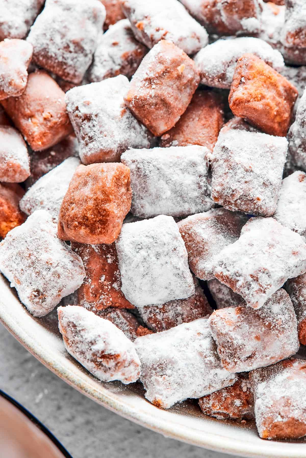 Close-up shot of puppy chow in a white bowl, to show the texture. Some pieces are fully coated in powdered sugar, while others are not.