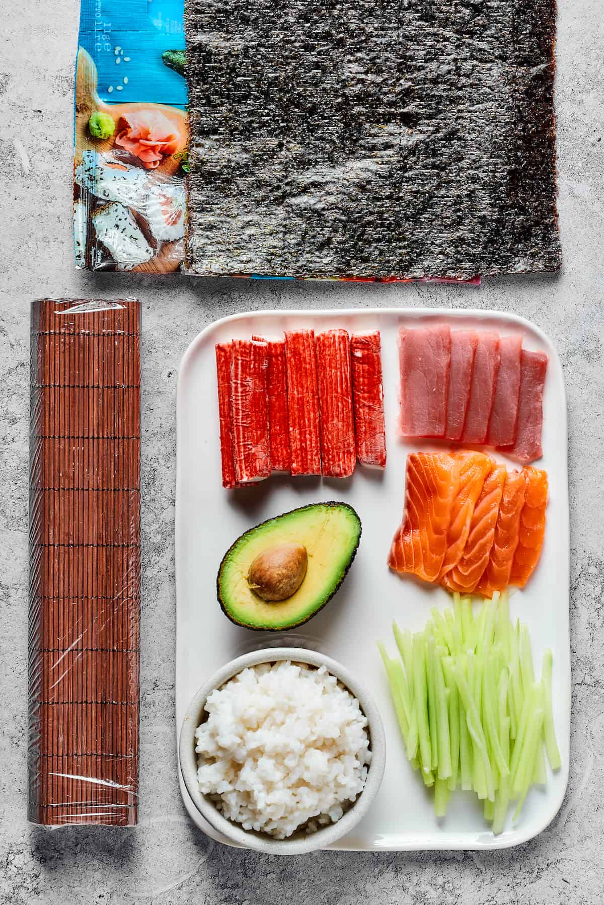 From top: Sheets of nori, a bamboo sushi mat, sticks of imitation crab, sliced raw tuna, an avocado, sliced raw salmon, prepared sushi rice, and sliced cucumber.
