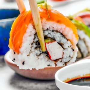 A bite of sushi held in a pair of chopsticks. A small plate of sushi and a container of soy sauce is in the background.