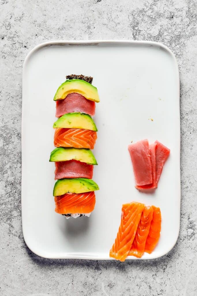 Overhead shot of a roll of sushi, topped with alternating slices of avocado, tuna, and salmon. Extra slices of tuna and salmon are on the platter.
