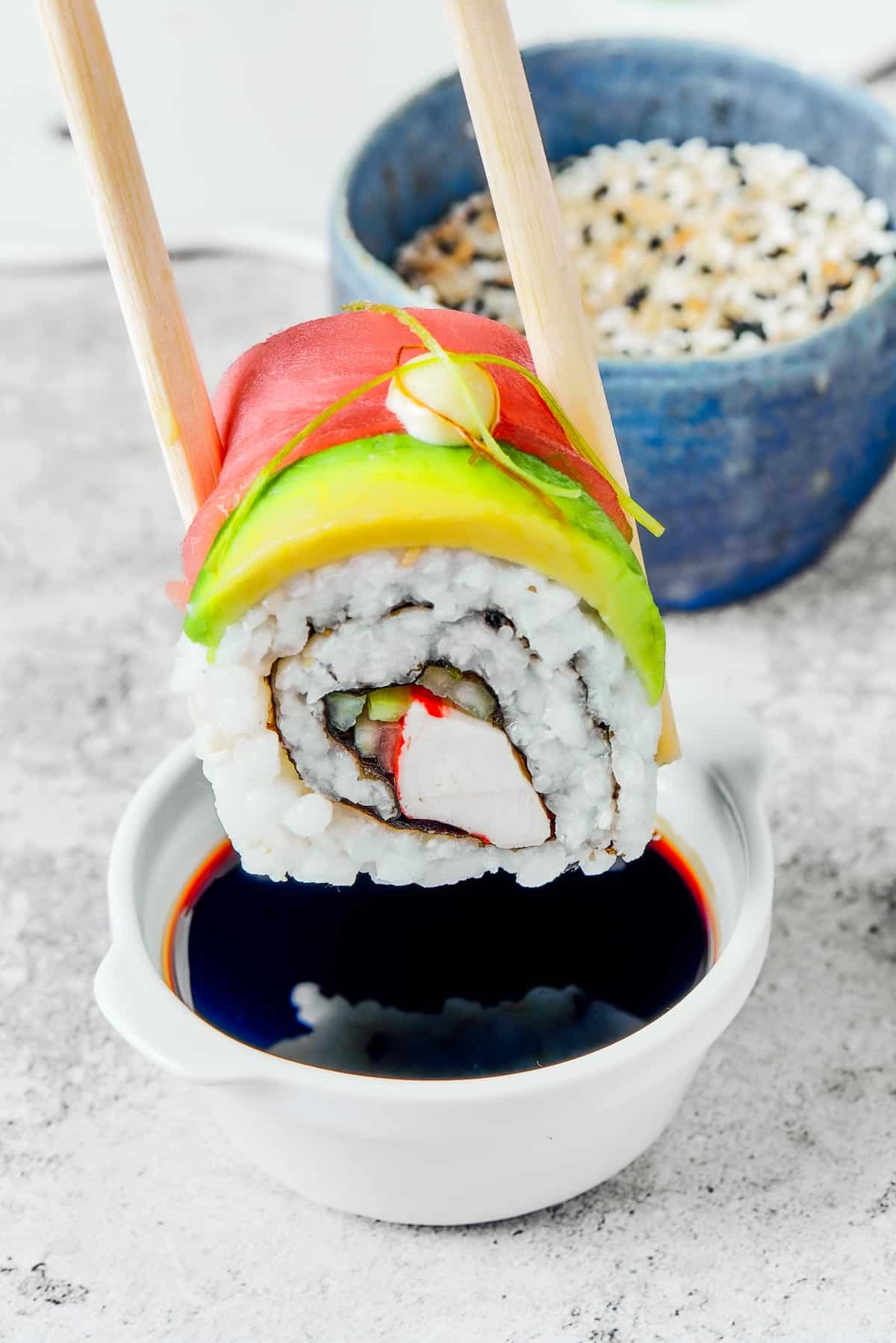 A bite of sushi held in a pair of chopsticks. A small plate of sushi and a container of soy sauce is in the background.