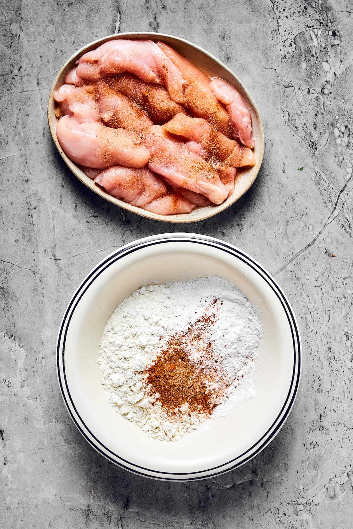 A dish of raw, seasoned chicken strips next to a bowl of seasoned flour.