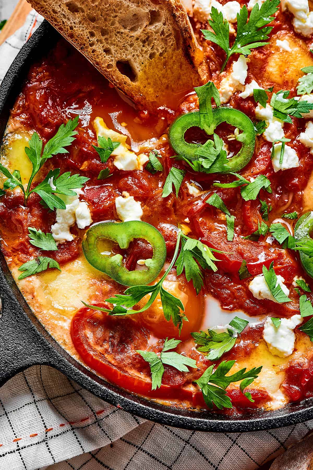 Shakshuka in a cast-iron skillet, with a baguette slice dipped in the sauce.