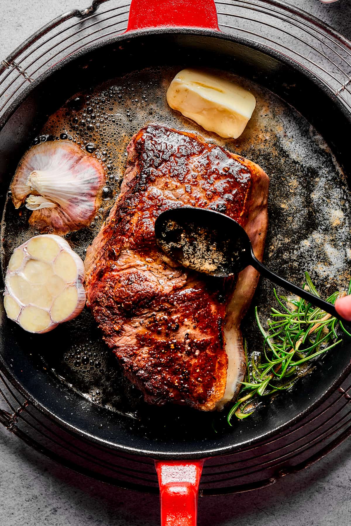 A person's hand basting pan juices over a steak, as the steak cooks in a skillet.