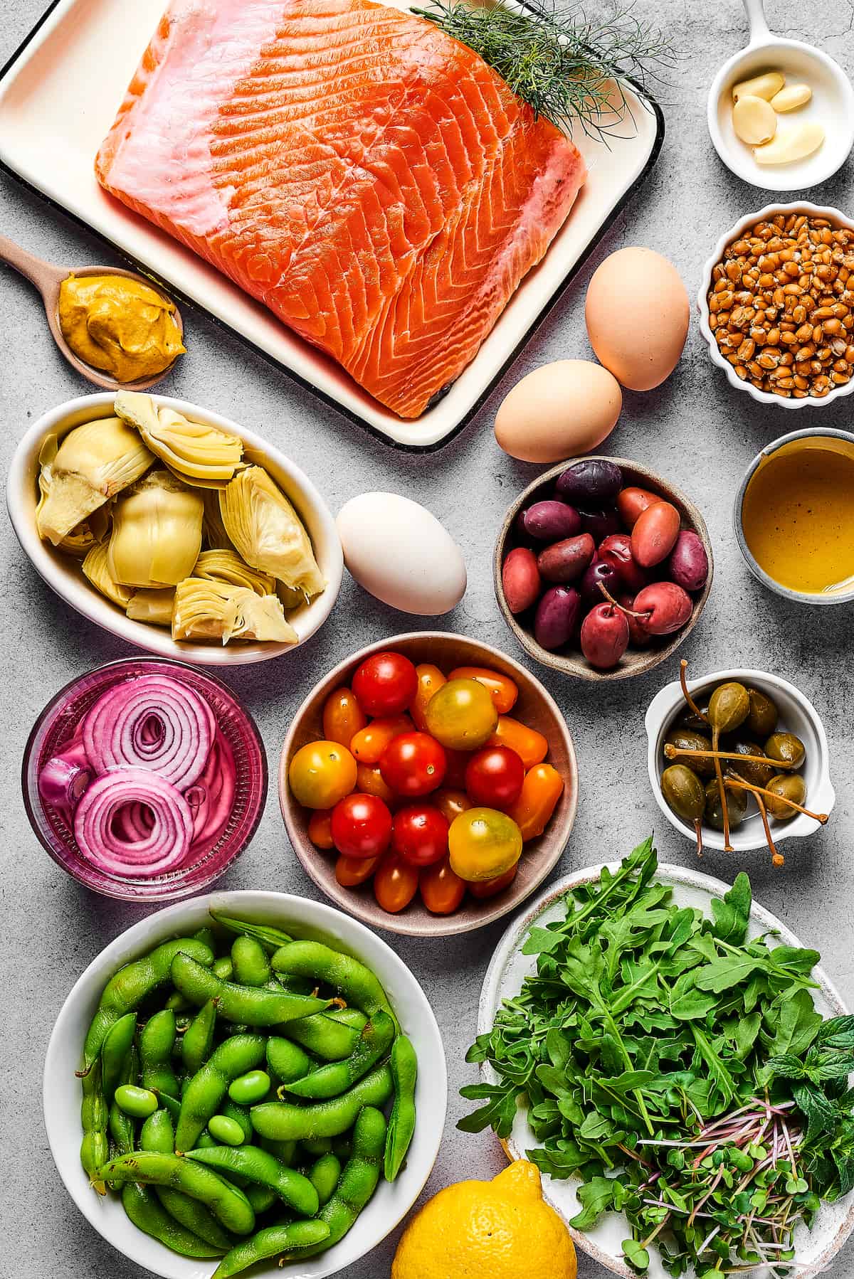 From top left: Salmon, garlic, dijon mustard, eggs, cooked grain, artichoke hearts, kalamata olives, olive oil, pickled red onions, tomatoes, capers, edamame, lemon, arugula.