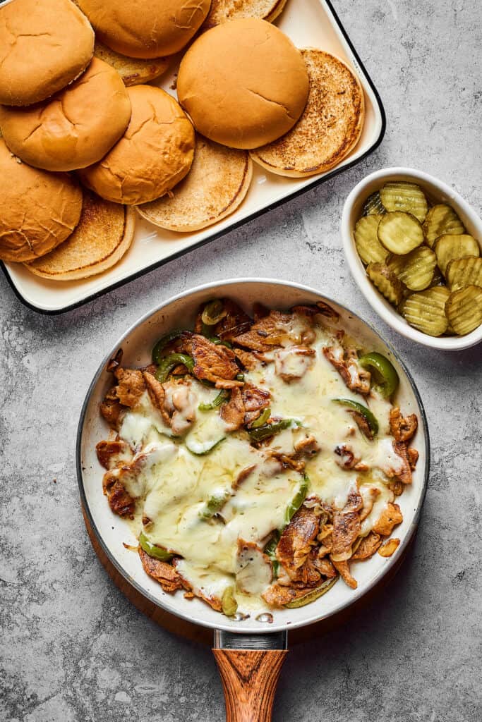 A tray of sandwich buns and a small dish of sliced dill pickles next to a skillet with cooked steak, onion, and bell pepper, topped with melted cheese.