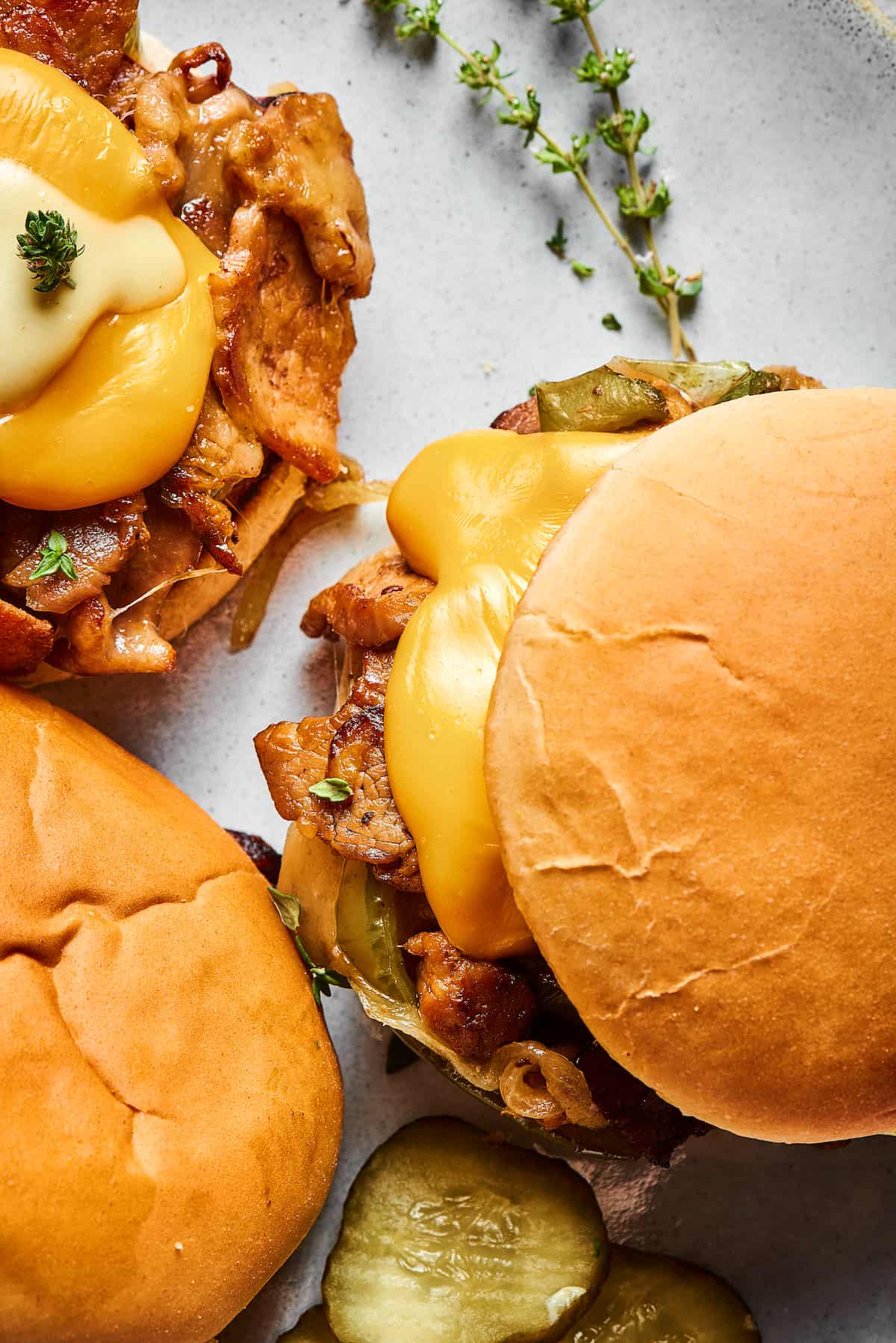 Close-up shot of three philly cheesesteak burgers on a platter with dill pickle chips and a sprig of thyme.